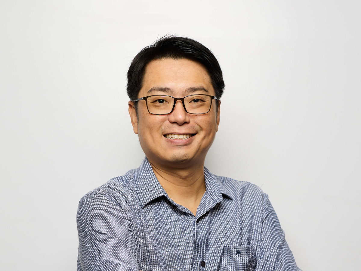 Mr Daniel Loh in glasses and a blue patterned shirt smiling with arms crossed against a light grey wall